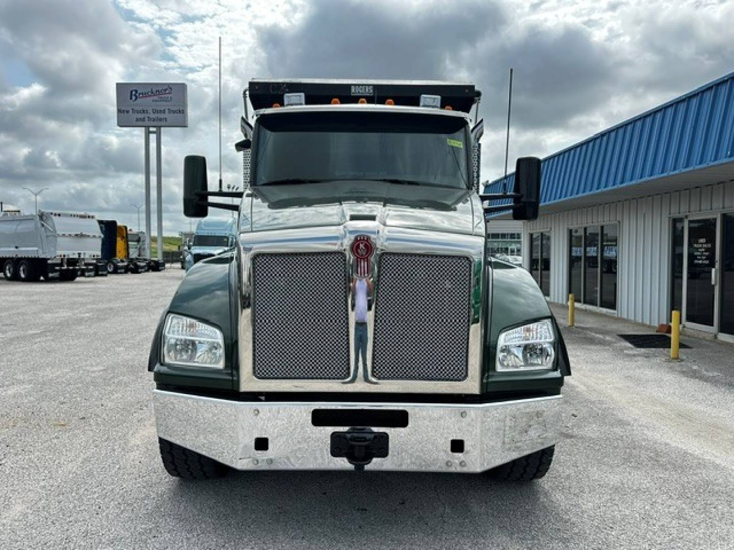 Kenworth T880 dump truck front view showing large chrome grille, rectangular headlights, and bumper; green body with plastic trim in a dealership lot.