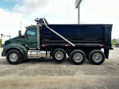 Kenworth T880 dump truck side view showing three-axle rear configuration, dark green cab, black dump body, chrome fuel tanks and exhaust, VIN plate not visible.