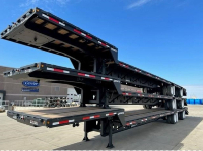 Dropdeck trailer by Demco showing multi-axle flatbed with hydraulic drop deck, black paint, reflective safety tape, stacked deck sections, landing gear, and truck yard in background.