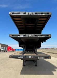 Demco dropdeck trailer in stacked position showing dual hinged decks and hydraulic lift mechanism with caution labels and frame cross members, blue sky background.