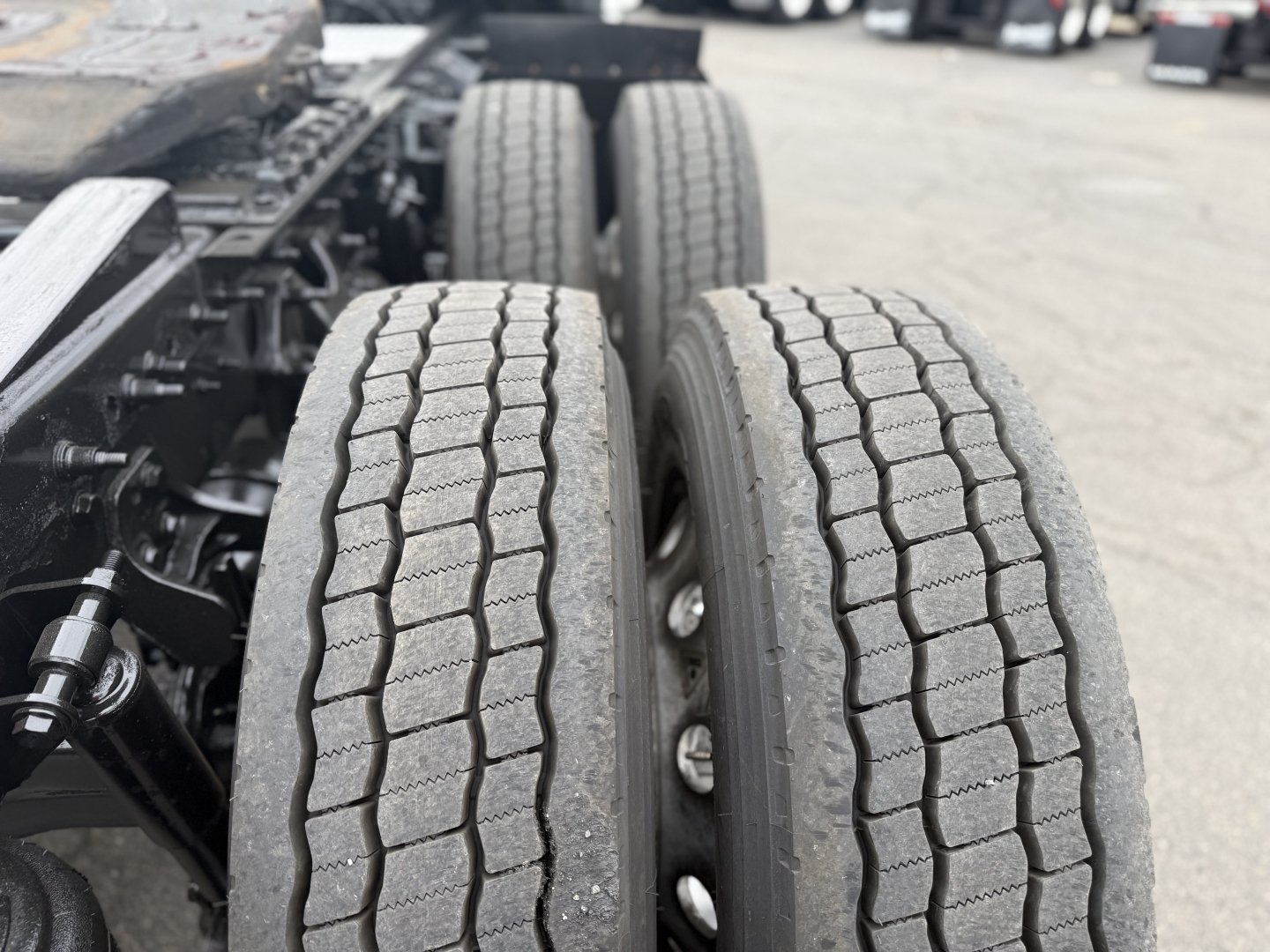 Dual rear tires on a tractor-trailer chassis with visible tread and wheel rims; portion of chassis and suspension components in the background.