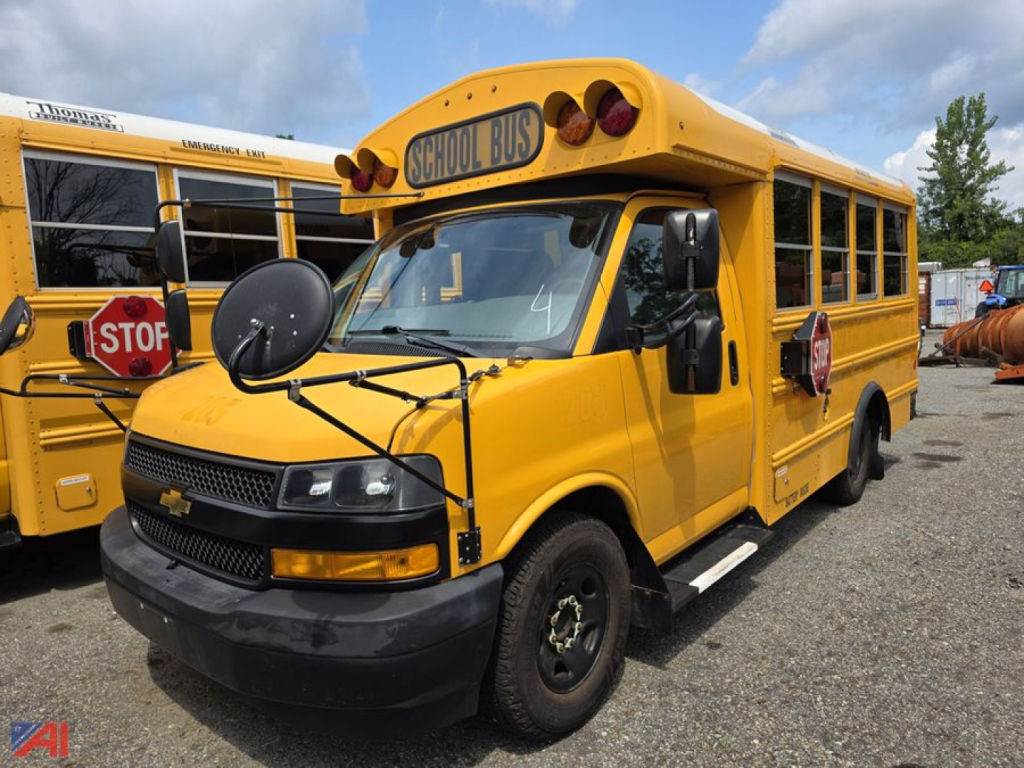 Yellow Chevrolet Express school bus with stop sign and school bus sign on front; side mirrors, black bumper, and roof lights visible; rear and another bus in background.