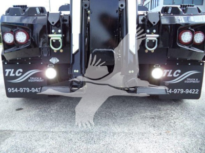 Truck winch platform rear view showing two TLC branding panels, taillights, hooks, and front of a heavy-duty trailer with hydraulic arms and safety latches.