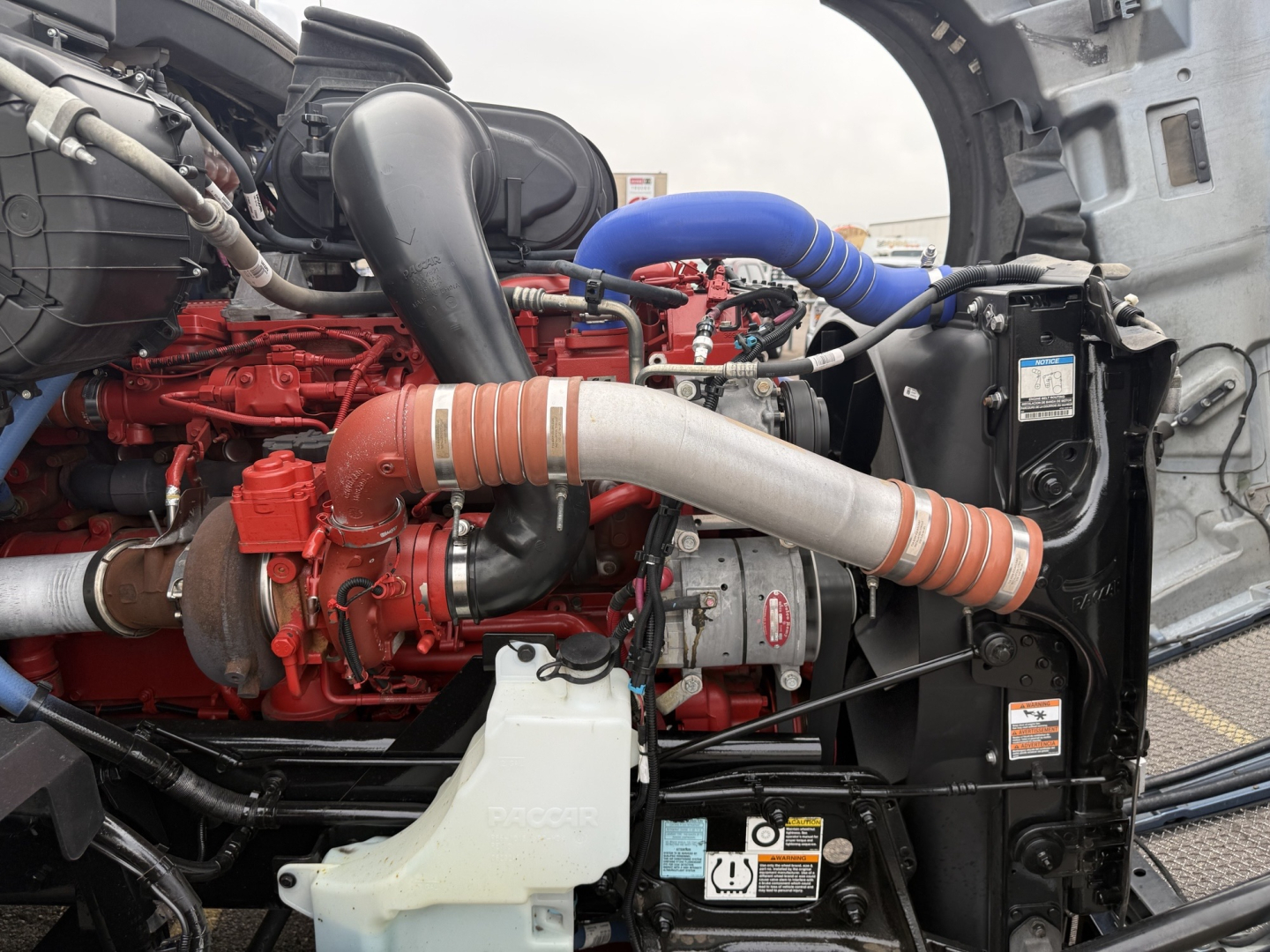 Kenworth T680 engine bay showing red engine block, blue silicone turbo hose, black intercooler piping, white coolant reservoir, and various labels.