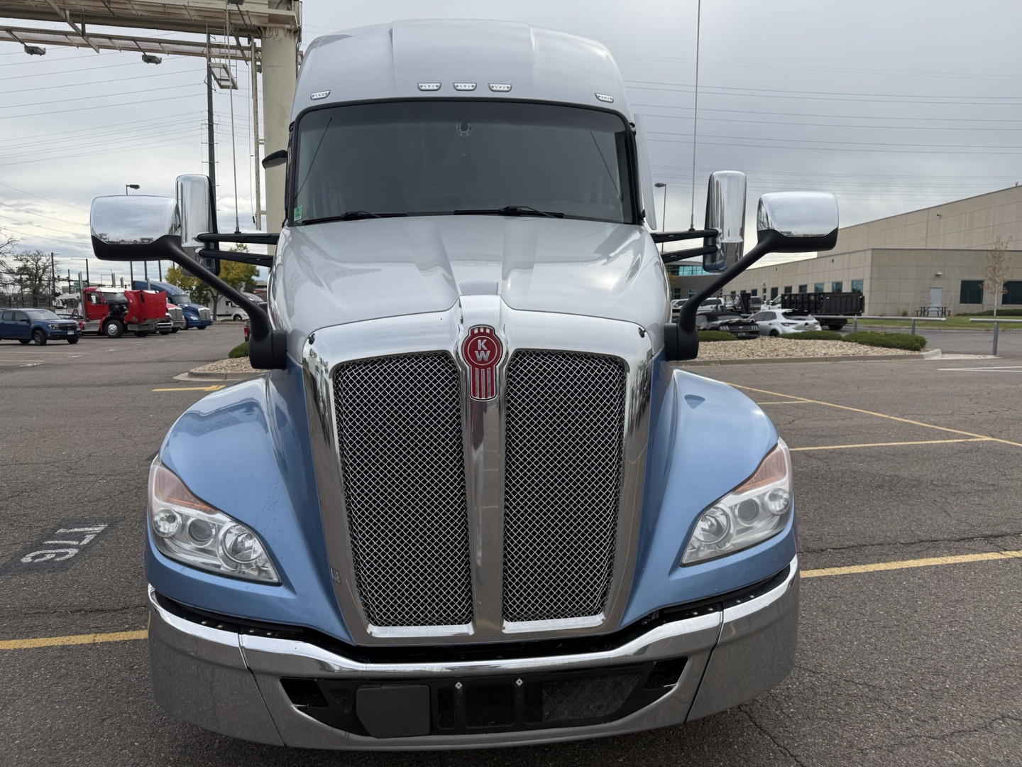 Kenworth T680 front view showing large dual grille with chrome surround, Kenworth emblem, blue fenders, silver bumper, side mirrors, and windshield. No visible text other than truck design.