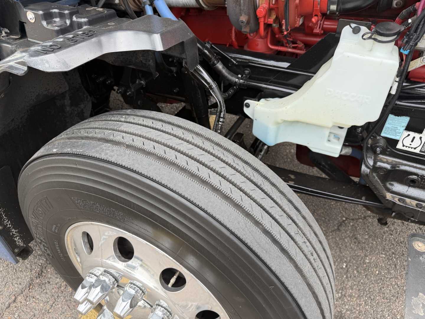 Kenworth T680 front wheel area showing dual front axle with large radial tires and white coolant reservoir; exposed engine components and frame.
