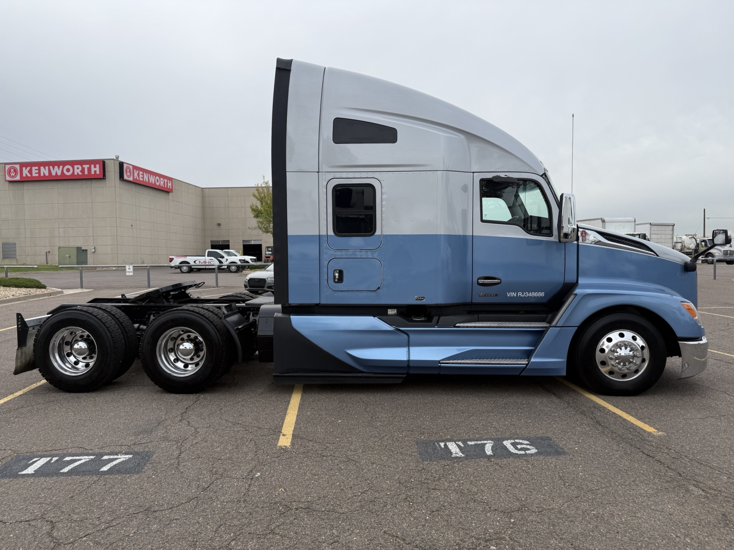 Kenworth T680 sleeper cab truck side profile showing blue lower body and silver upper cab, dual rear axles, with wheel rims and Kenworth branding visible in background.