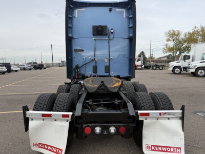Kenworth T680 rear view showing fifth wheel, dual rear axles, and suspension; blue cab section with wiring and components mounted; Kenworth mud flaps and taillights visible.