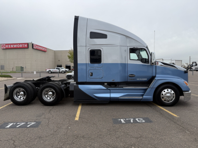 Kenworth T680 sleeper cab truck side profile showing blue lower body and silver upper cab, dual rear axles, with wheel rims and Kenworth branding visible in background.