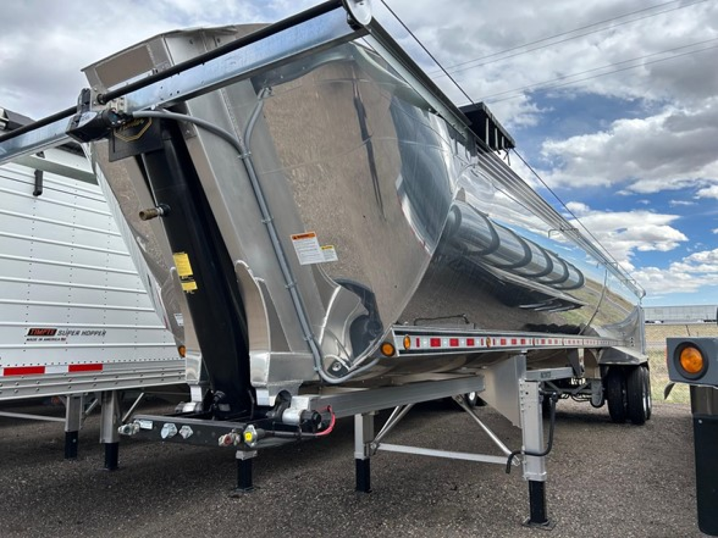 Dump trailer showing polished steel hopper with front tongue and adjustable legs, side reflective stripes, and hydraulic hoist hardware visible.