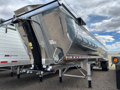Dump trailer showing polished steel hopper with front tongue and adjustable legs, side reflective stripes, and hydraulic hoist hardware visible.