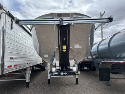 Trailer hitch in front of a dump trailer with a raised support arm and hydraulic system, visible from the rear of the trailer showing the hitch, frame, and safety chains.