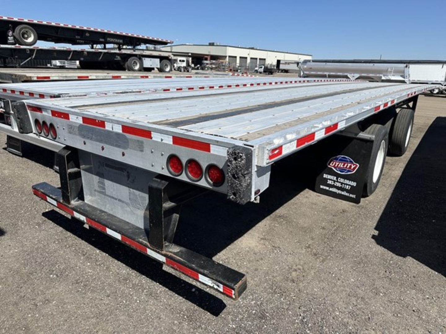Flatbed trailer rear and side with metal deck, red and white reflective tape, multiple taillights, spare tire, and mudflaps showing ‘Utility Trailer’ mudflap branding.