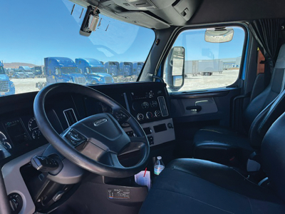 Cab interior of a Freightliner Cascadia truck showing steering wheel, dashboard, and driver seat; daytime outside with other trucks visible.