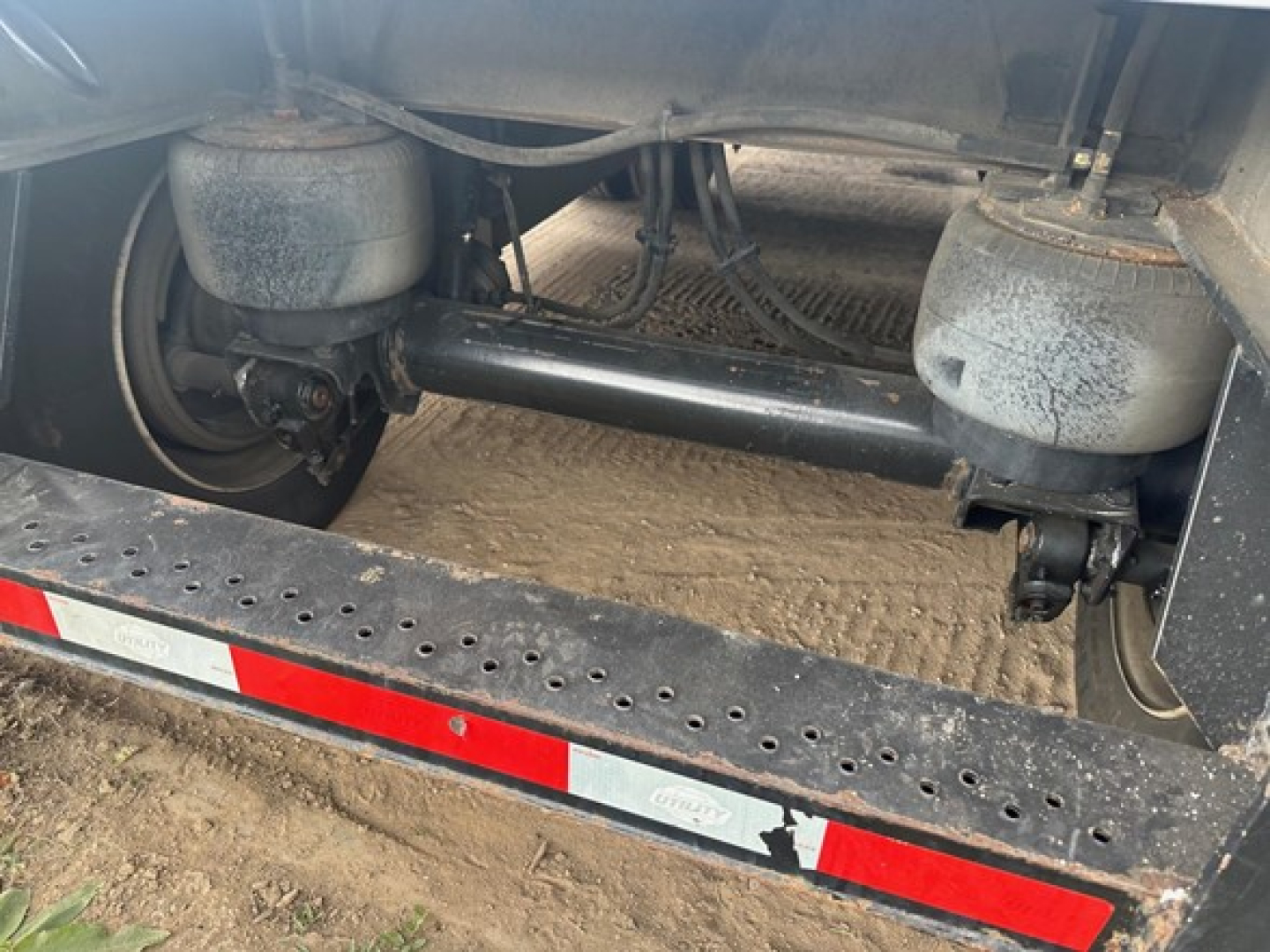 Trailer undercarriage showing dual air springs and solid axle with a black crossmember and frame rails; side step with reflective red/white tape; dirt ground beneath.