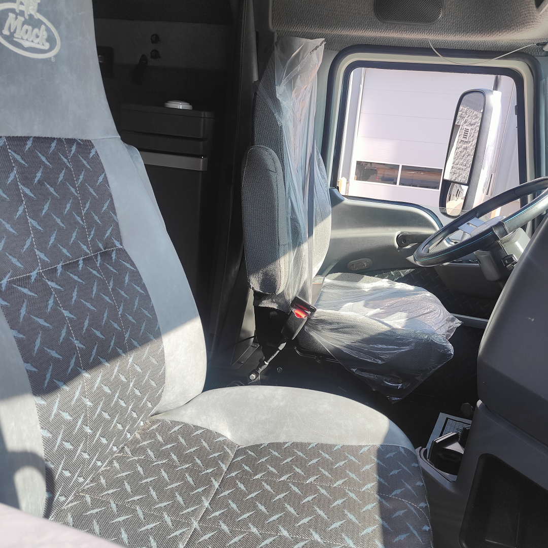 Truck cab interior of a 2016 Mack CXU613 showing driver seat and steering wheel with plastic seat covers, visible side window, doorway, and dashboard features.