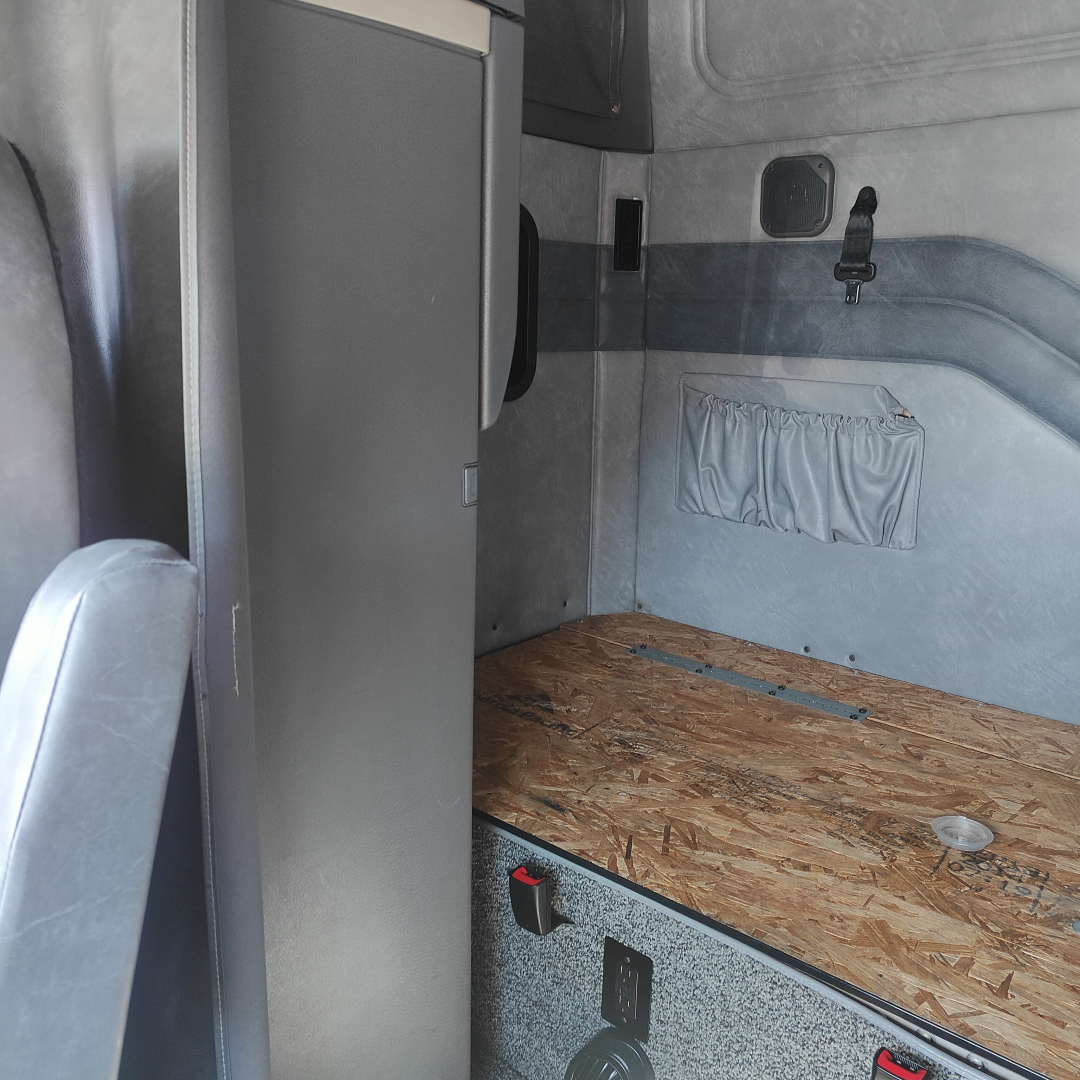 Cab interior of a Mack CXU613 sleeper cab showing a raised bed area with plywood floor, small storage pocket on wall, and a gray cabinet.