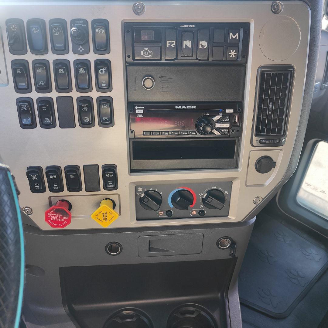 Dashboard of a Mack CXU613 truck showing control switches, a Mack audio unit, climate dials, and warning labels.