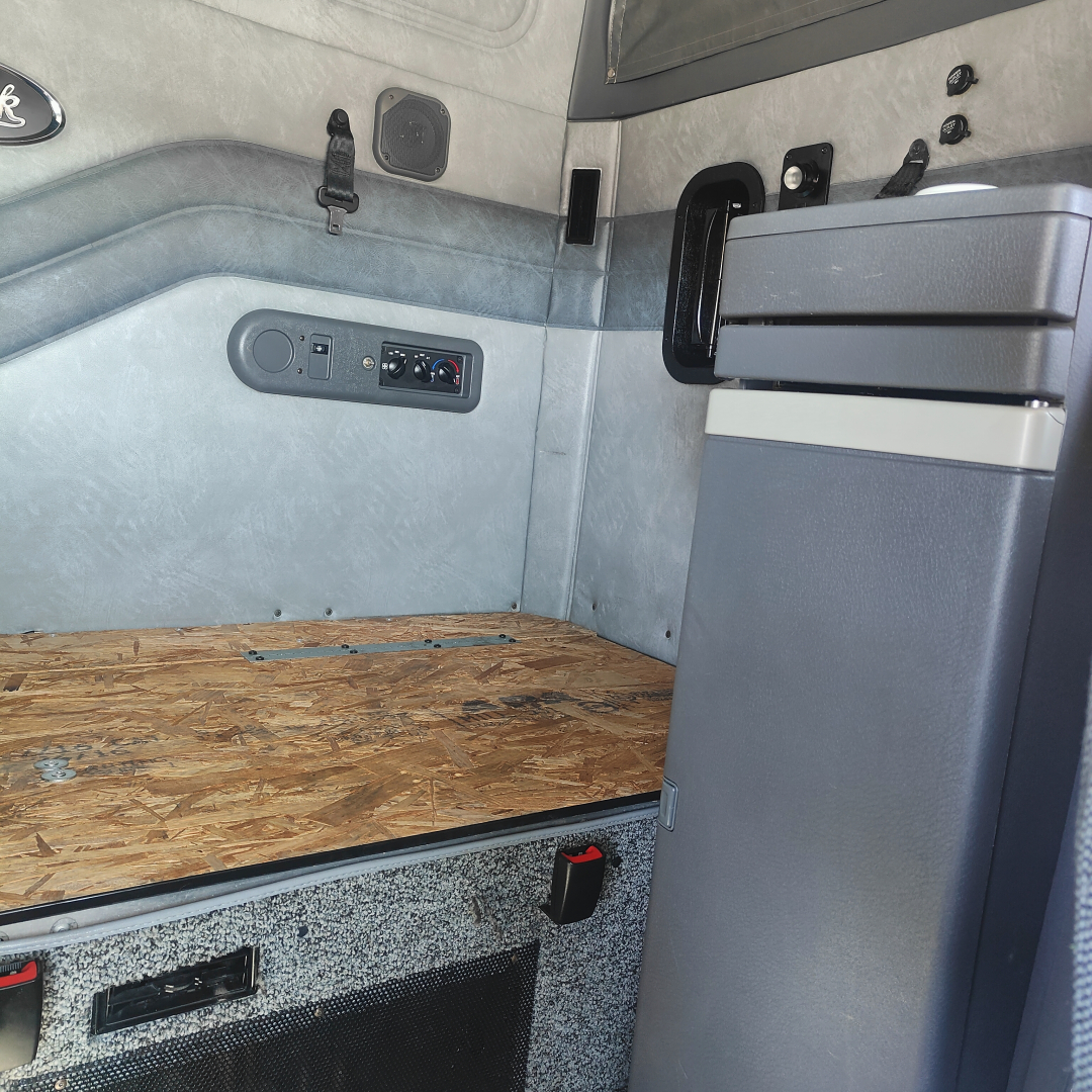 Truck cab interior of a 2016 Mack CXU613 sleeper showing side wall with storage, controls, and a raised plywood floor in the cargo area.