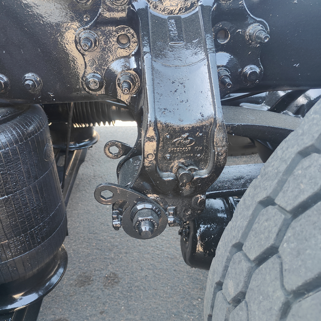 Truck rear suspension and leaf spring mounting hardware; close-up of bracket and bolts near a large tire, painted black with oily texture.