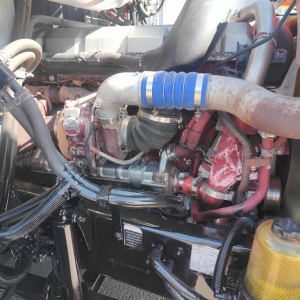 Engine bay view of a 2016 Mack CXU613 showing turbo intake hose, aluminum piping, red painted components, hoses, and a yellow coolant reservoir cap.