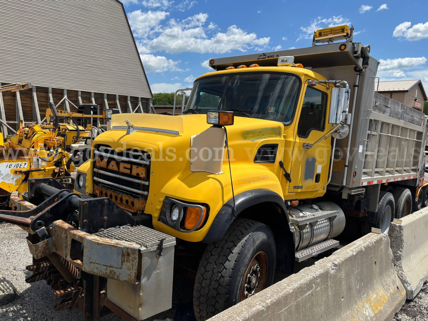 Mack CV713 dump truck in yellow with a front grille and daytime running lights, viewed from the driver side, showing the dump bed and front bumper.