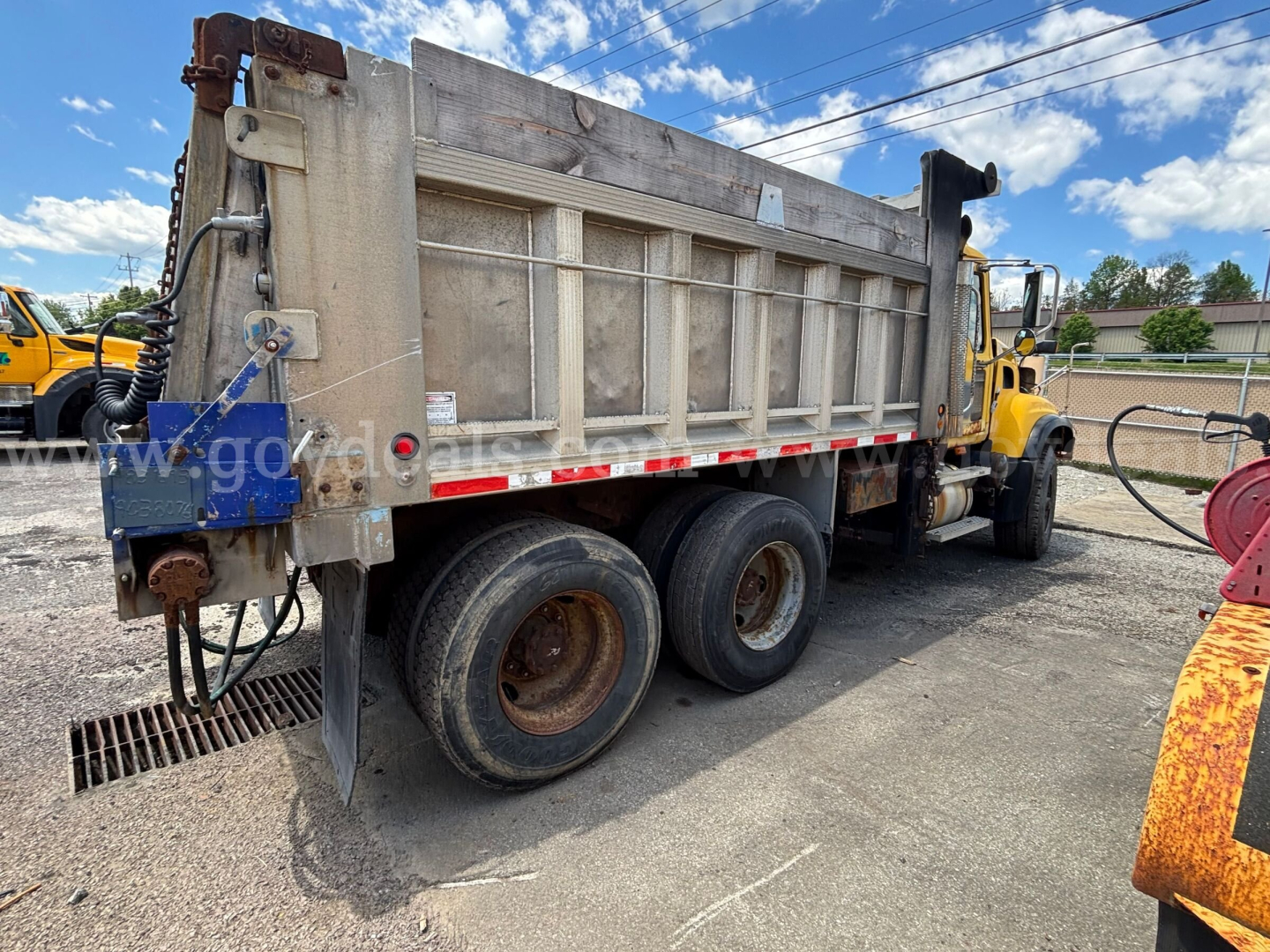 Dump truck side rear view showing three rear axles on a yellow Mack CV713 with metal dump bed and hydraulic components.