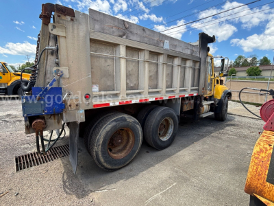 Dump truck side rear view showing three rear axles on a yellow Mack CV713 with metal dump bed and hydraulic components.