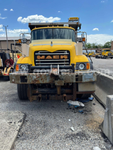 Mack CV713 dump truck front view with yellow cab and grilles, visible bumper, headlights, and rusted front reinforcement plate.