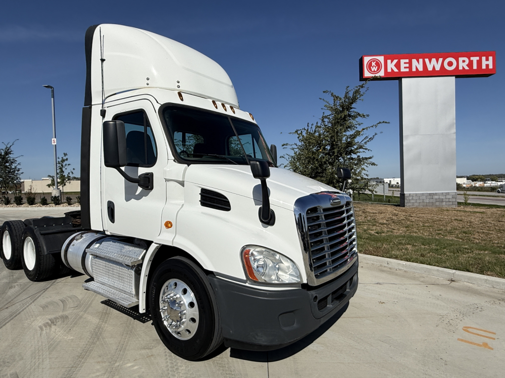 Truck cab with white Freightliner CA11364DC semi-truck, Kenworth sign in background. Visible text includes  Kenworth sign and Freightliner styling.