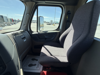 Truck cab interior of a Freightliner CA11364DC showing steering wheel, dashboard, and driver's seat; side window and exterior lot visible through windshield.