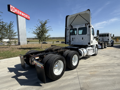 Freightliner CA11364DC tractor in a truck lot showing rear triple-axle drive and raised cab. Visible text on sign reads KENWORTH; truck lacks load.
