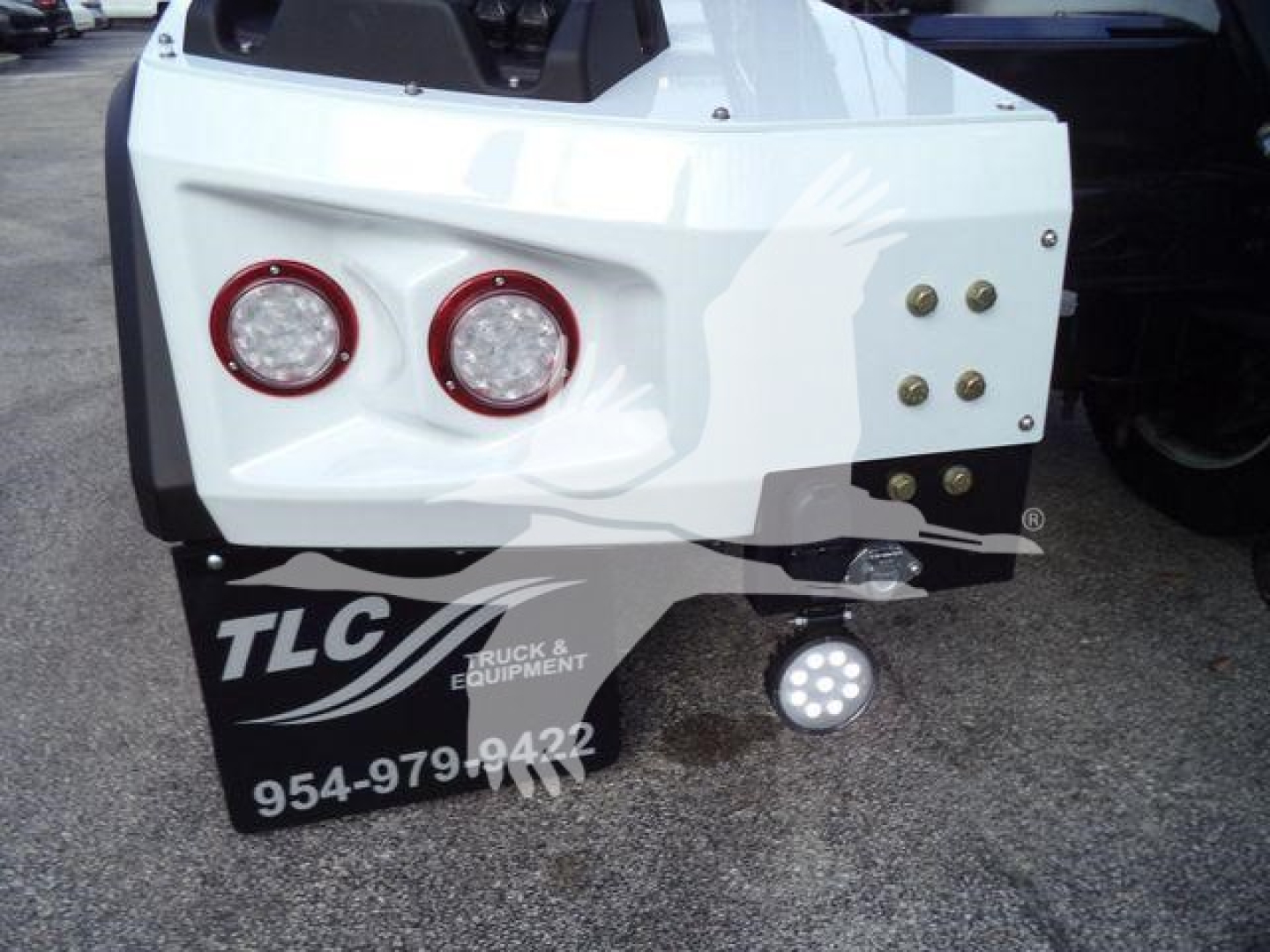 Rear bumper of a white Chevrolet Silverado 6500HD winch truck showing two round red taillights, a set of bolts on the right, a TCL Truck & Equipment mudflap, and a round LED light on a hitch mount.