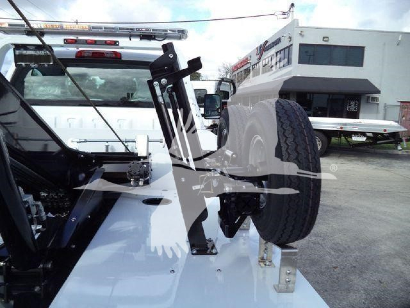 Truck bed showing a spare tire mounted and hydraulic winch assembly with metal brackets and hydraulic components on a white Chevrolet Silverado 6500HD chassis; loading dock and building in background.