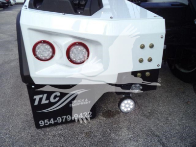 Rear bumper of a white Chevrolet Silverado 6500HD winch truck showing two round red taillights, a set of bolts on the right, a TCL Truck & Equipment mudflap, and a round LED light on a hitch mount.