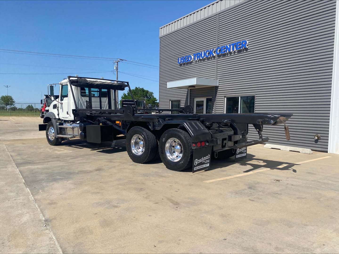 Mack GU533 roll-off truck on lot, side view showing chassis and dual rear axles with flatbed hooklift. Building with sign in background.