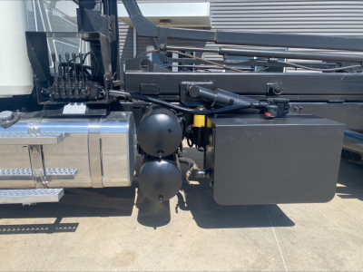 Trucks side view showing hydraulic hoses, twin black spherical tanks, and a black rectangular toolbox mounted near the frame of a Mack GU533 roll-off truck.