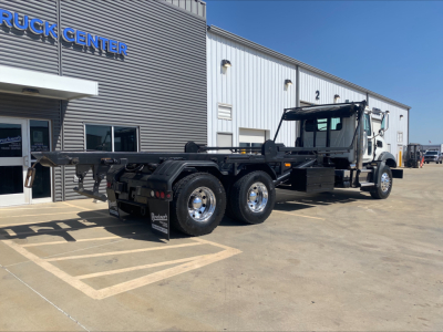 Rear and side view of a Mack GU533 roll-off truck with chassis exposed, showing six tires, hydraulic lines, and a Mack dealer building in the background. Visible text: 'TRUCK CENTER' on building sign.