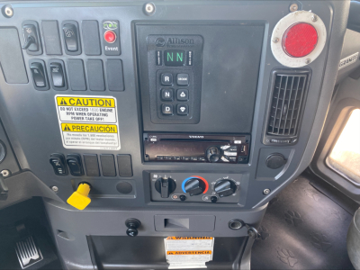 Interior dash of a 2018 Mack GU533 showing control panel with gear selector, warning labels, climate controls, and radio. Visible text includes caution labels and digital gear indicator.