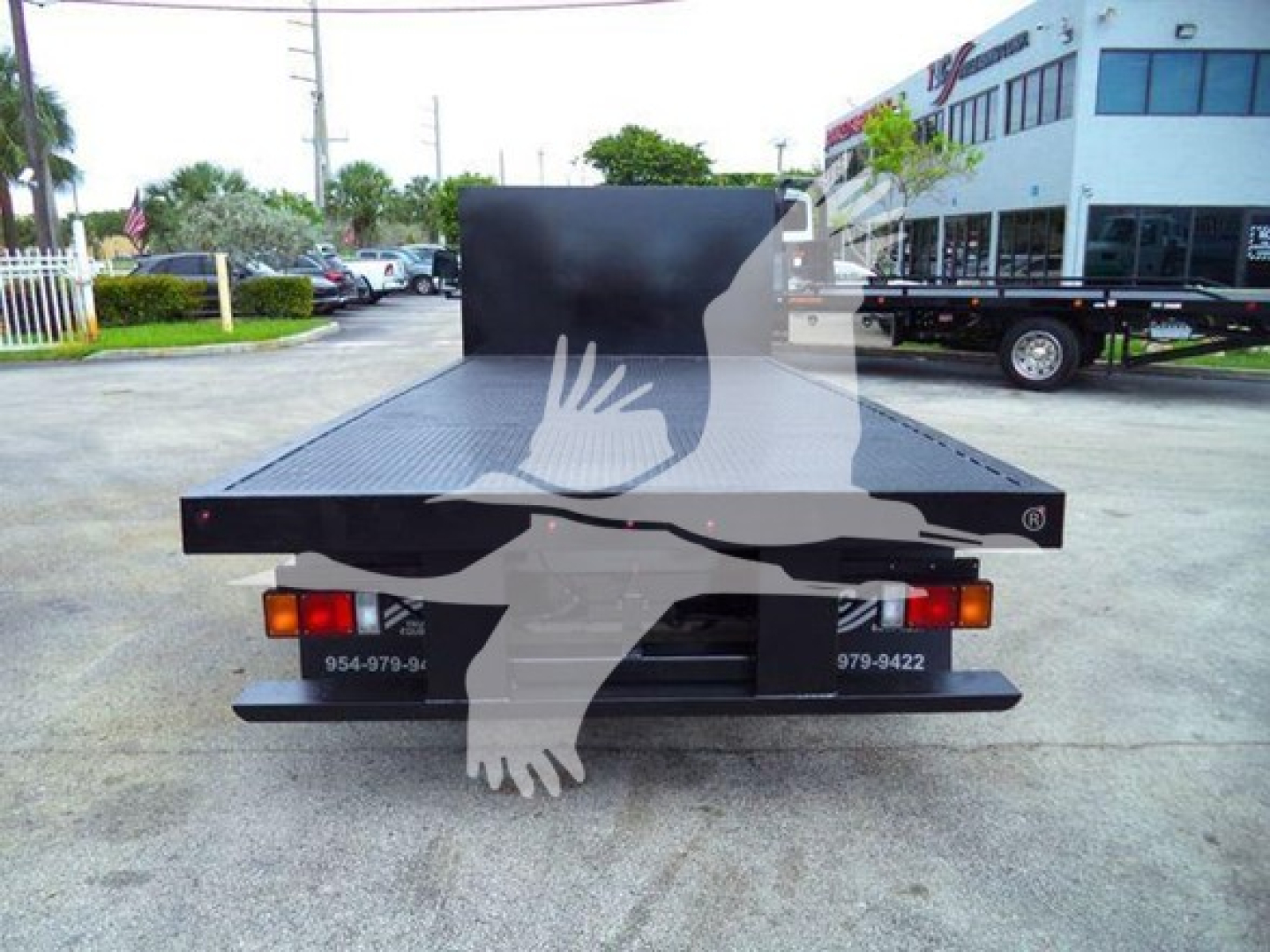 Flatbed tow truck rear view showing a black steel platform and tail lights, with phone numbers on the bumper and a tall bulkhead at the front of the bed.