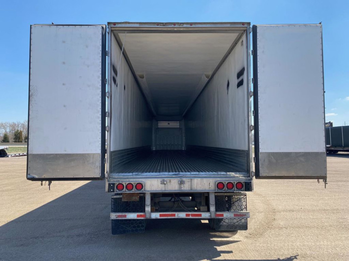 Trailer rear view with open doors showing empty refrigerated cargo area; metal floor with ribbed texture; rear lights and bumper visible.