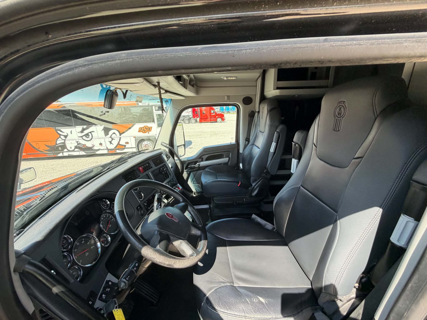 Cab interior of a 2022 Kenworth T680, steering wheel, dashboard gauges, and two black leather seats visible in a sleeper cab.