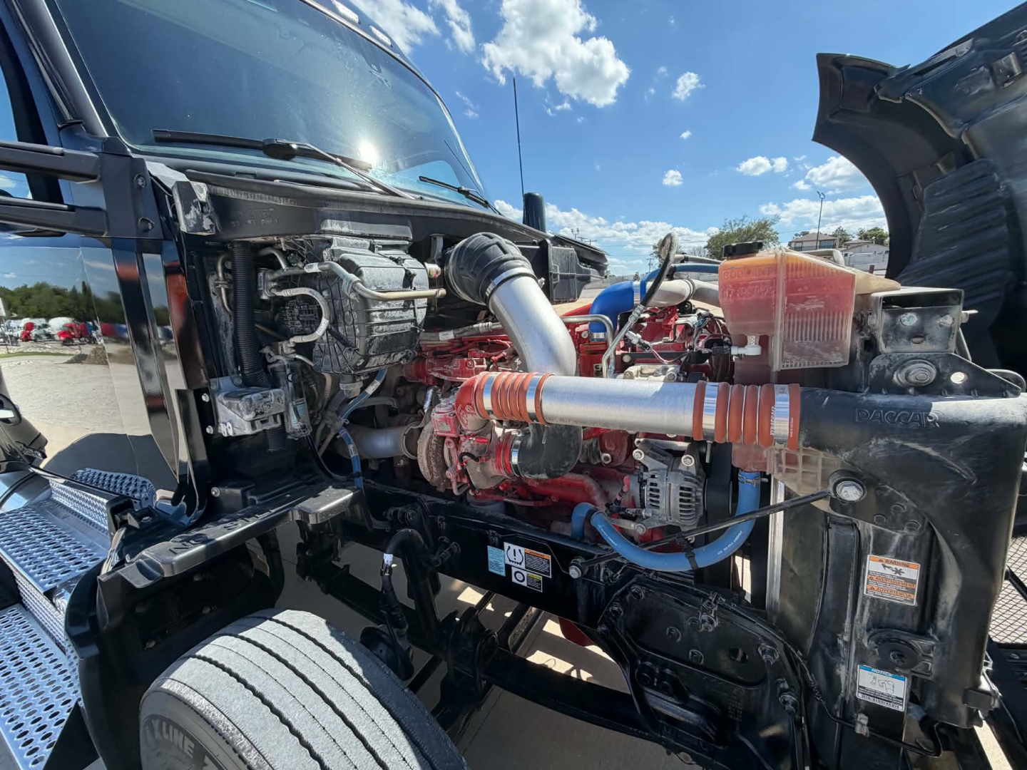 Kenworth T680 engine bay showing intercooler piping and turbo, coolant reservoir, and engine components in a daytime setting.