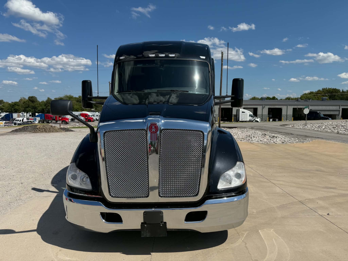 Kenworth T680 front view with chrome grille and bumper, big black mirrors, blue cab. No visible text on vehicle badges from distance.