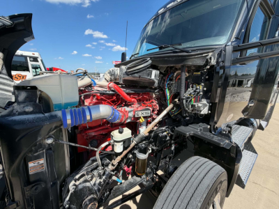 Kenworth T680 engine bay showing red engine with blue hose, interconnecting wiring, and exposed components under open hood.