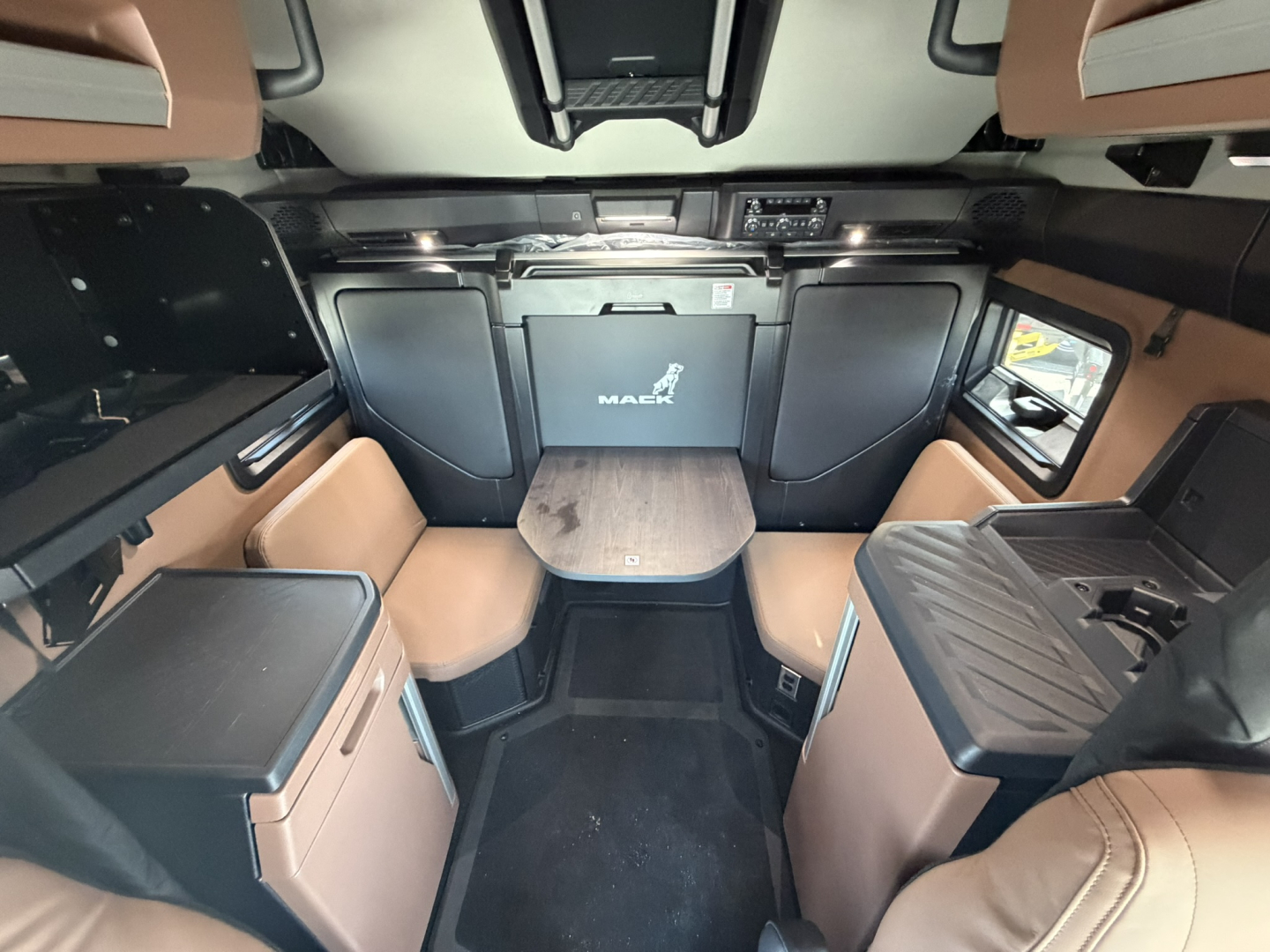Mack PR64T76H sleeper cab interior showing front dash with driver seat area, overhead storage, and central table; tan and black interior with cabinetry and seating.