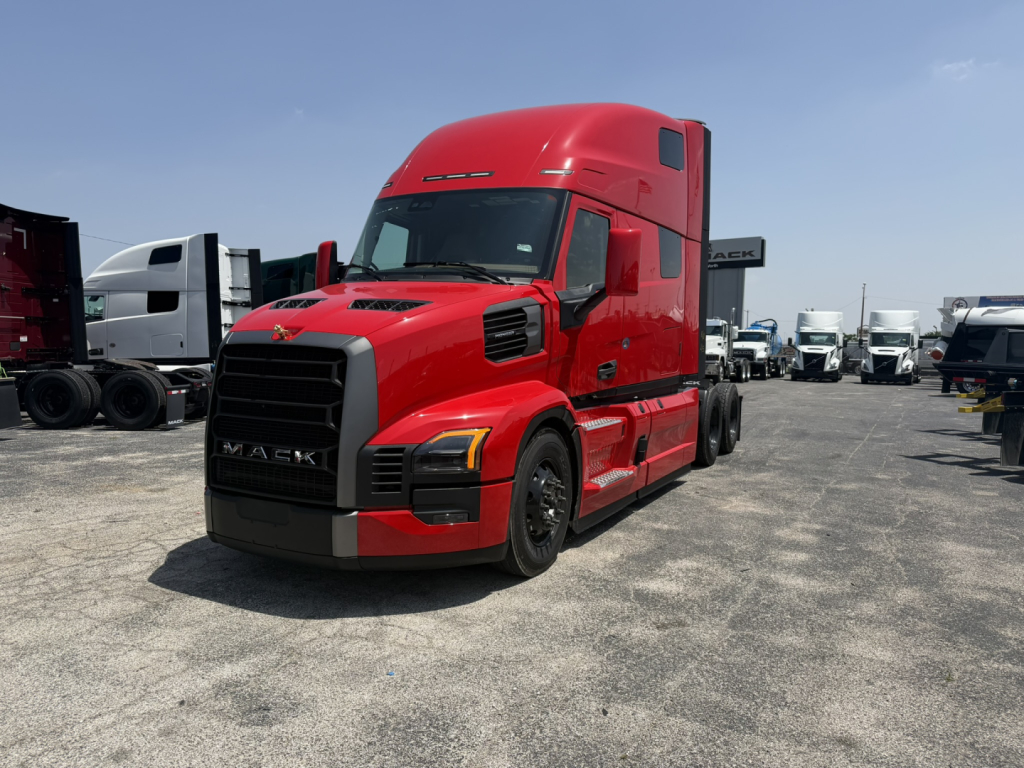 Red Mack PR64T76H tractor in a lot with other trucks; front grille with Mack emblem, headlights, and aerodynamic hood. Clear sky.