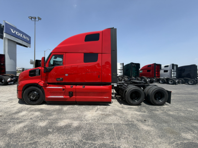Truck side profile showing red 2027 Mack PR64T76H sleeper cab tractor at a dealership lot with other trucks in background.