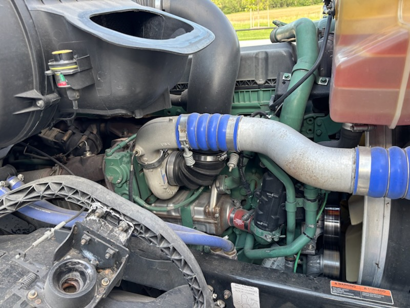 Engine bay with turbo piping and blue silicone hoses visible in a Volvo VNL780. Green engine block, black intake, and various hoses and belts.