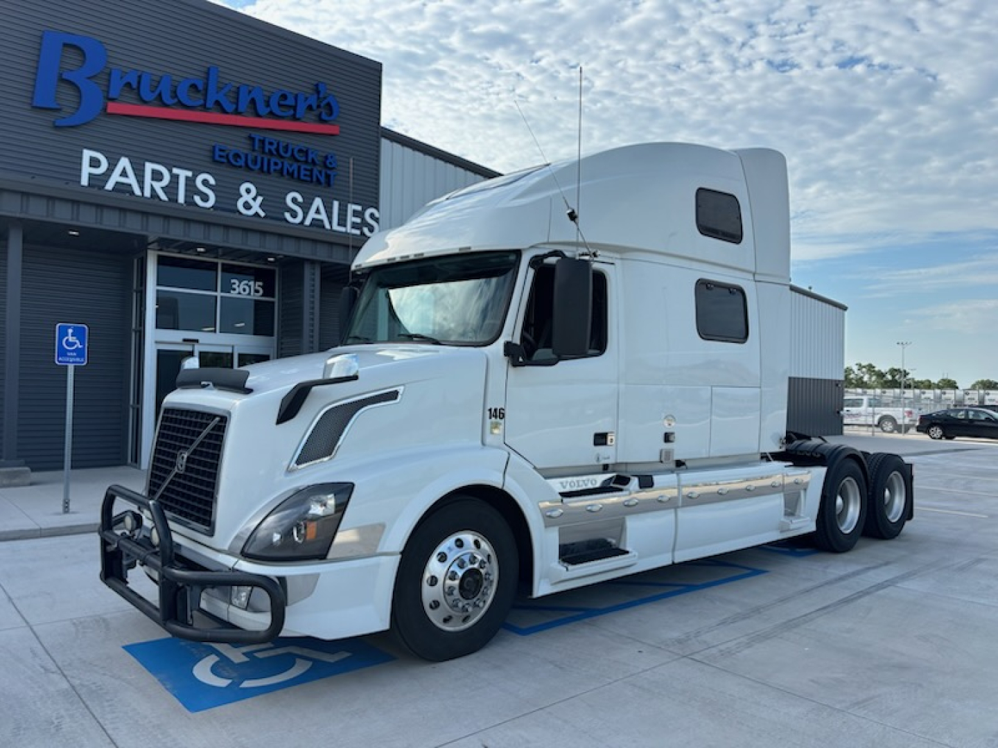 Volvo VNL780 truck in white, sleeper cab, front view parked at dealer lot with ‘Parts & Sales’ building in background.
